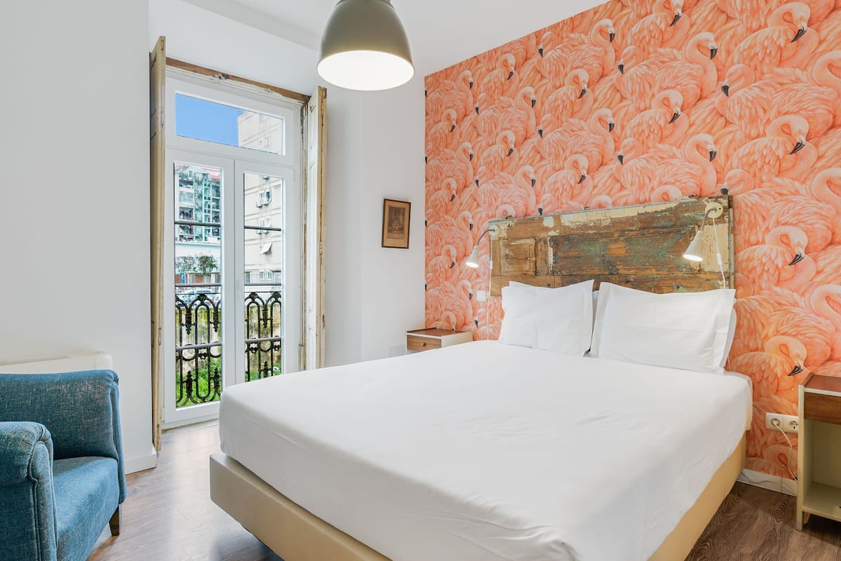 A bedroom with a large bed, a wooden headboard, a nightstand, a blue chair, and a window with a view of buildings.