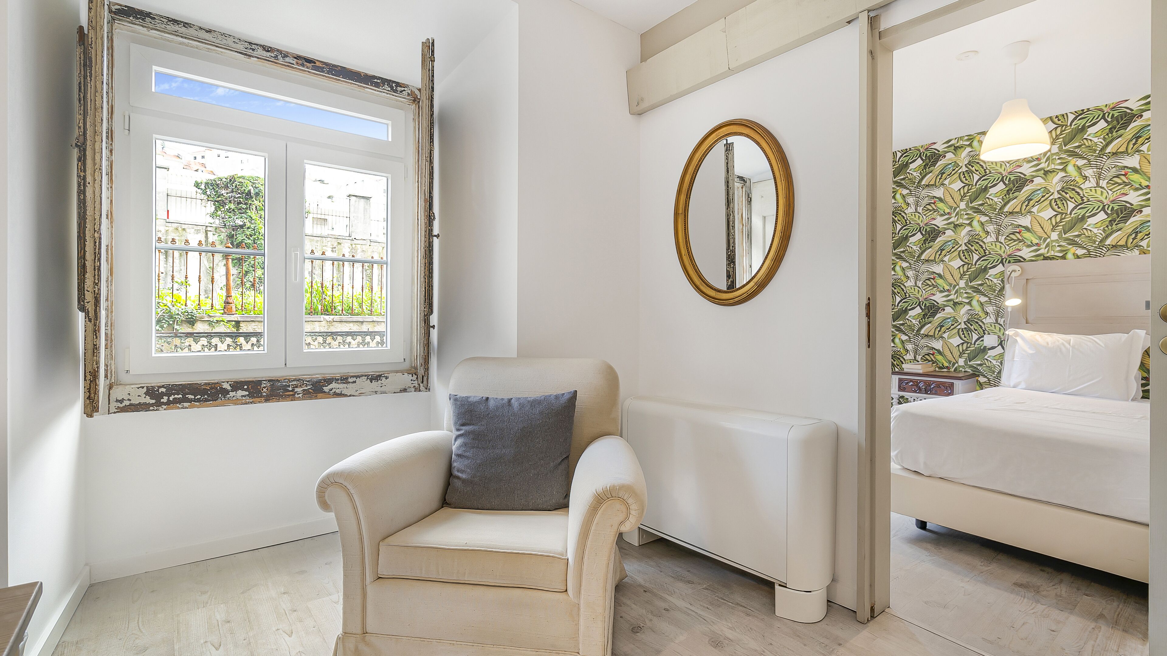 Una habitación con un sillón blanco, una cama con ropa blanca, un espejo grande y una ventana con vistas a edificios y zonas verdes.