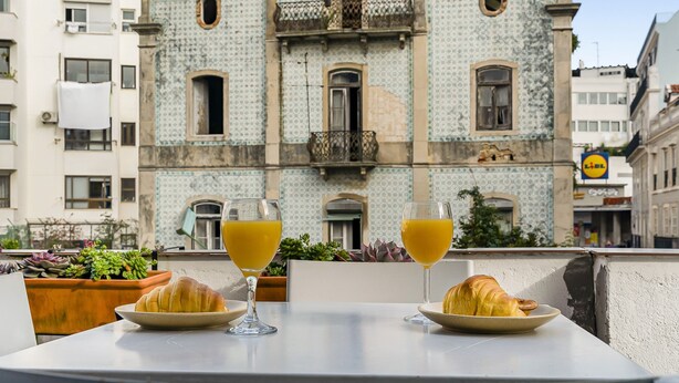Un tavolo con due croissant e due bicchieri di succo d'arancia, sullo sfondo di un edificio storico con piastrelle decorate e balconi.