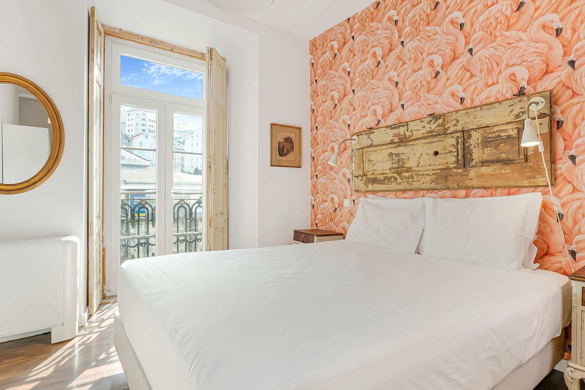 A bedroom with a large bed, a wooden headboard, a bedside table with a lamp, and a window with a view of buildings.
