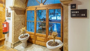 Ingresso di un hotel con una porta in legno, due piante in vaso, un estintore e un cartello che indica le camere 105-106.