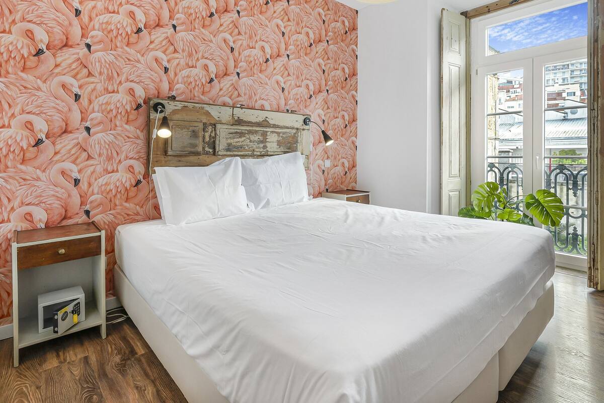 A bedroom with a bed, nightstands, a wall clock, and a window with a view of buildings and a balcony.