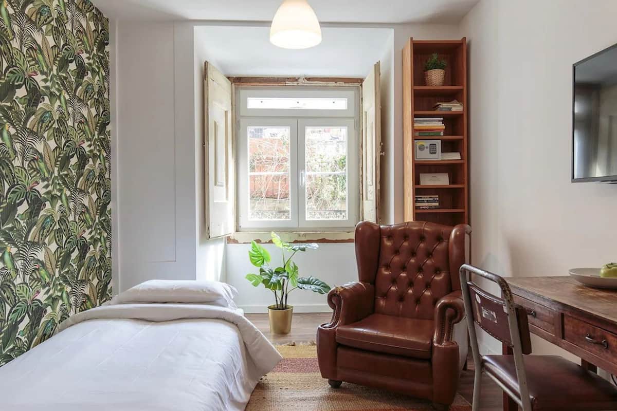 A bedroom with a bed, a wooden chair, a desk, a bookshelf, a television, and a window with a view of the outside.