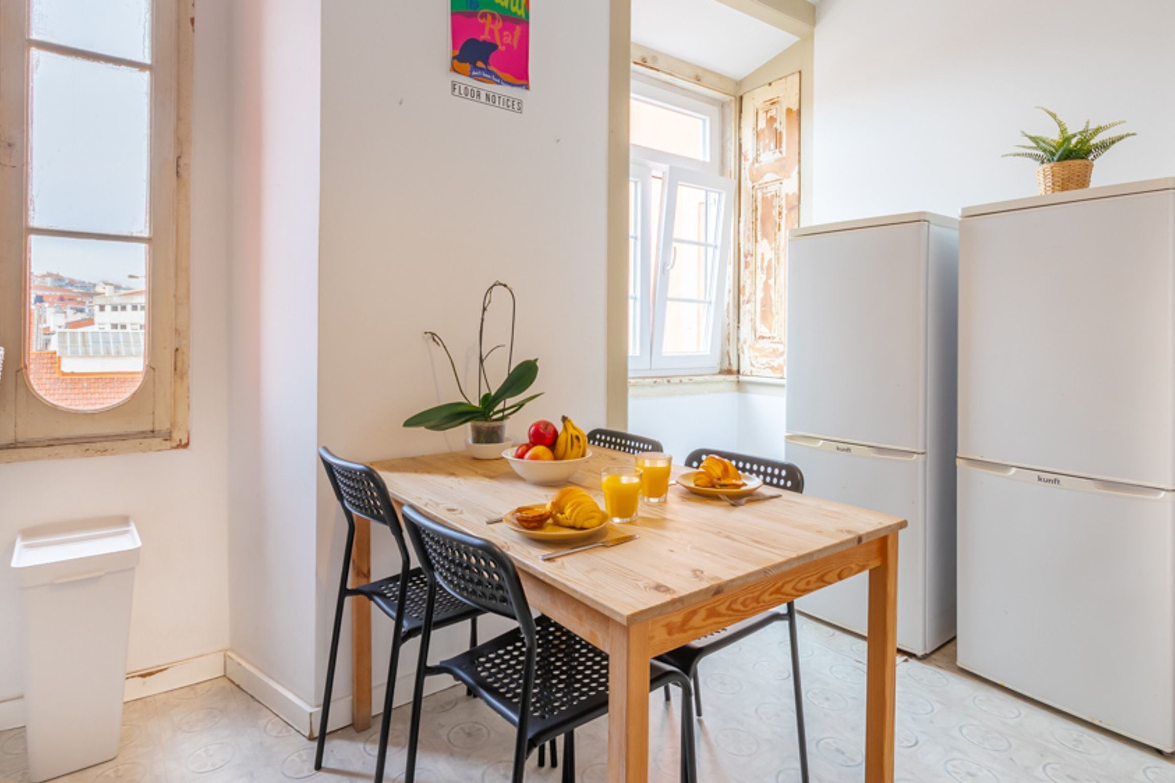 Una piccola cucina con un tavolo di legno, sedie nere, un frigorifero e una finestra vista edifici.