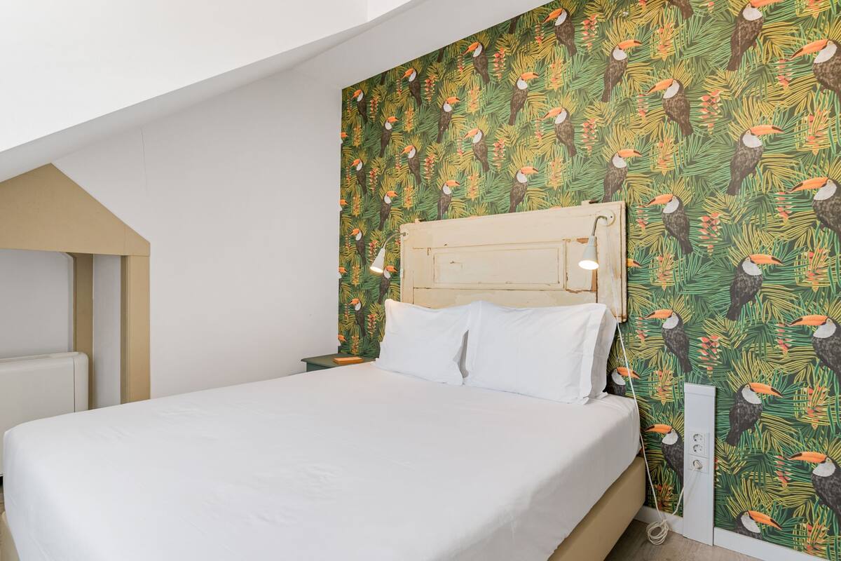 A bed with white linens, a wooden headboard, and a nightstand with a lamp. The wall features a tropical leaf and toucan wallpaper.