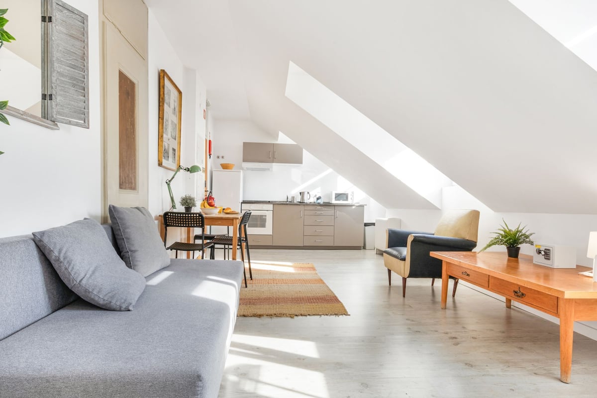 A modern loft apartment with a loft bed, a dining area, a kitchen, and a living room with a gray sofa and wooden desk.
