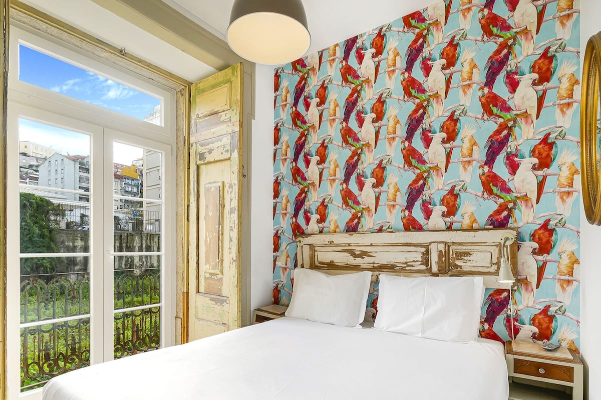 A bedroom with a large bed, a nightstand, a lamp, and a window with a view of buildings and greenery.