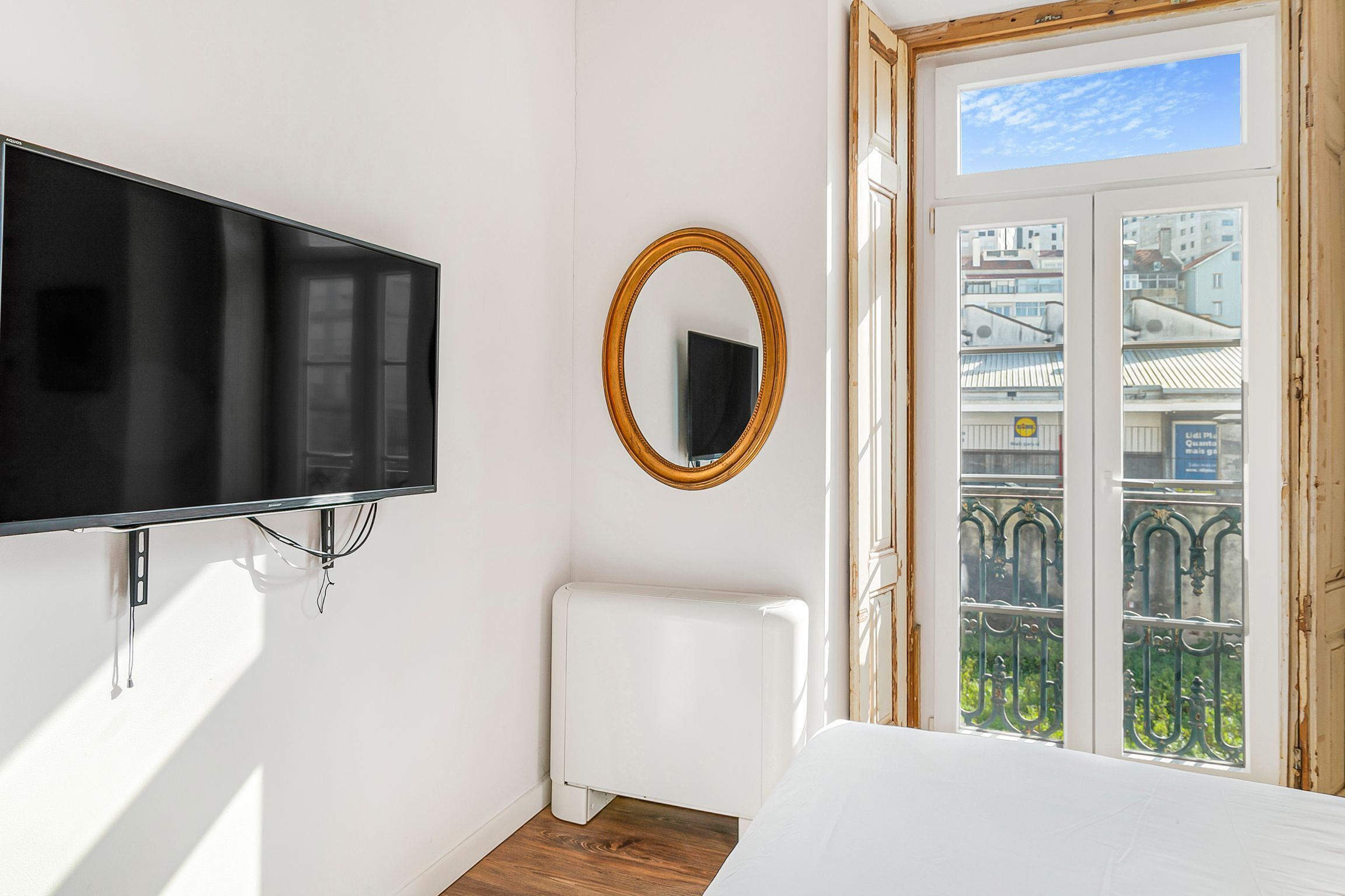 Un dormitorio con televisor montado en la pared, radiador blanco y ventana con vista a edificios y un balcón.