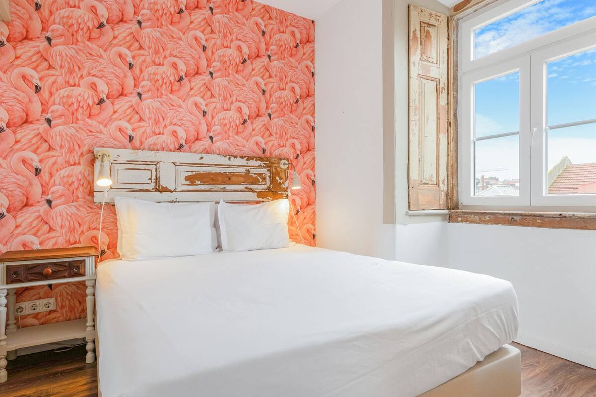 A bedroom with a white bed, a nightstand, a wall with a pink flamingo pattern, and a window with a view of buildings.