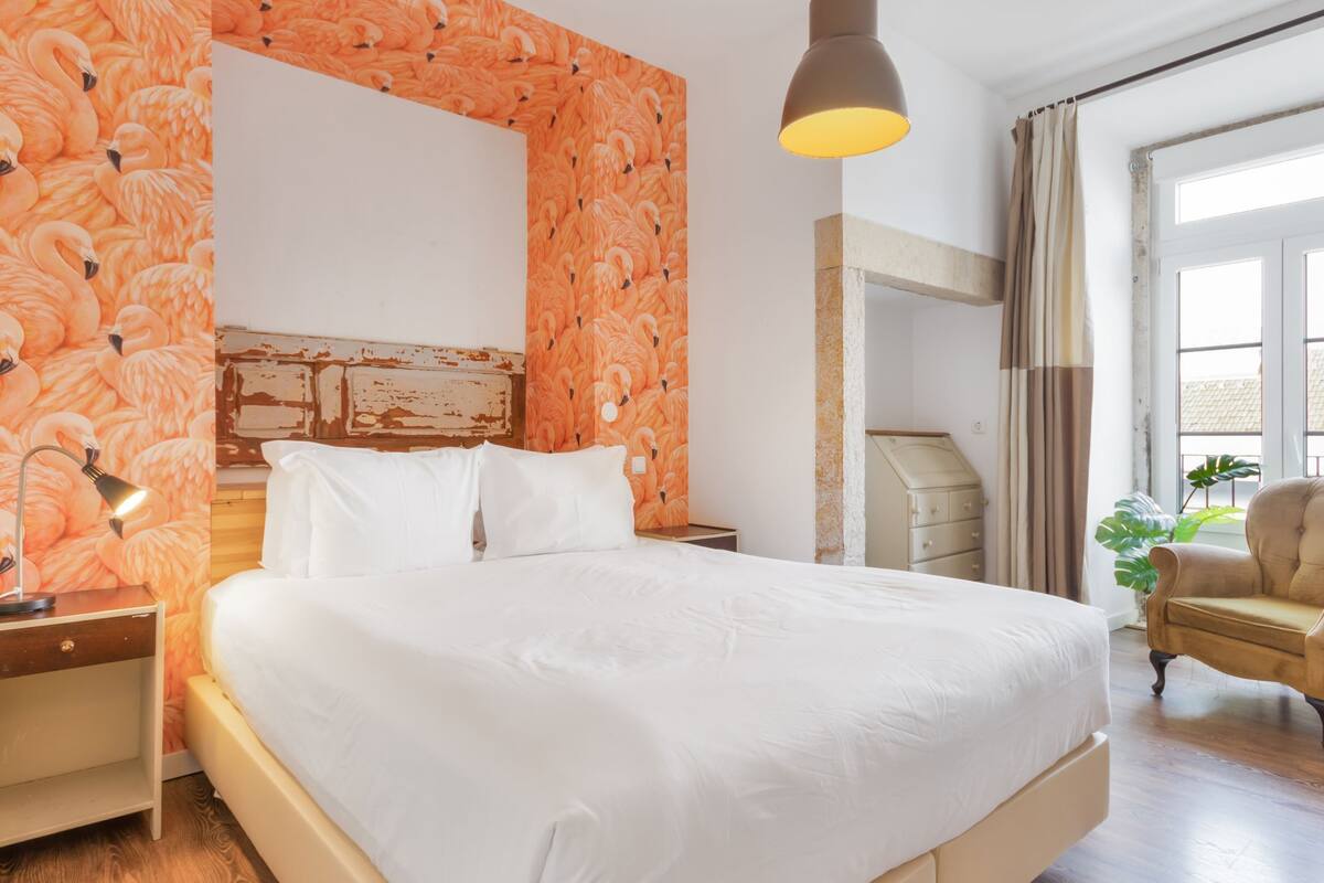 A hotel room with a large bed, a nightstand, a lamp, a chair, and a window with curtains.