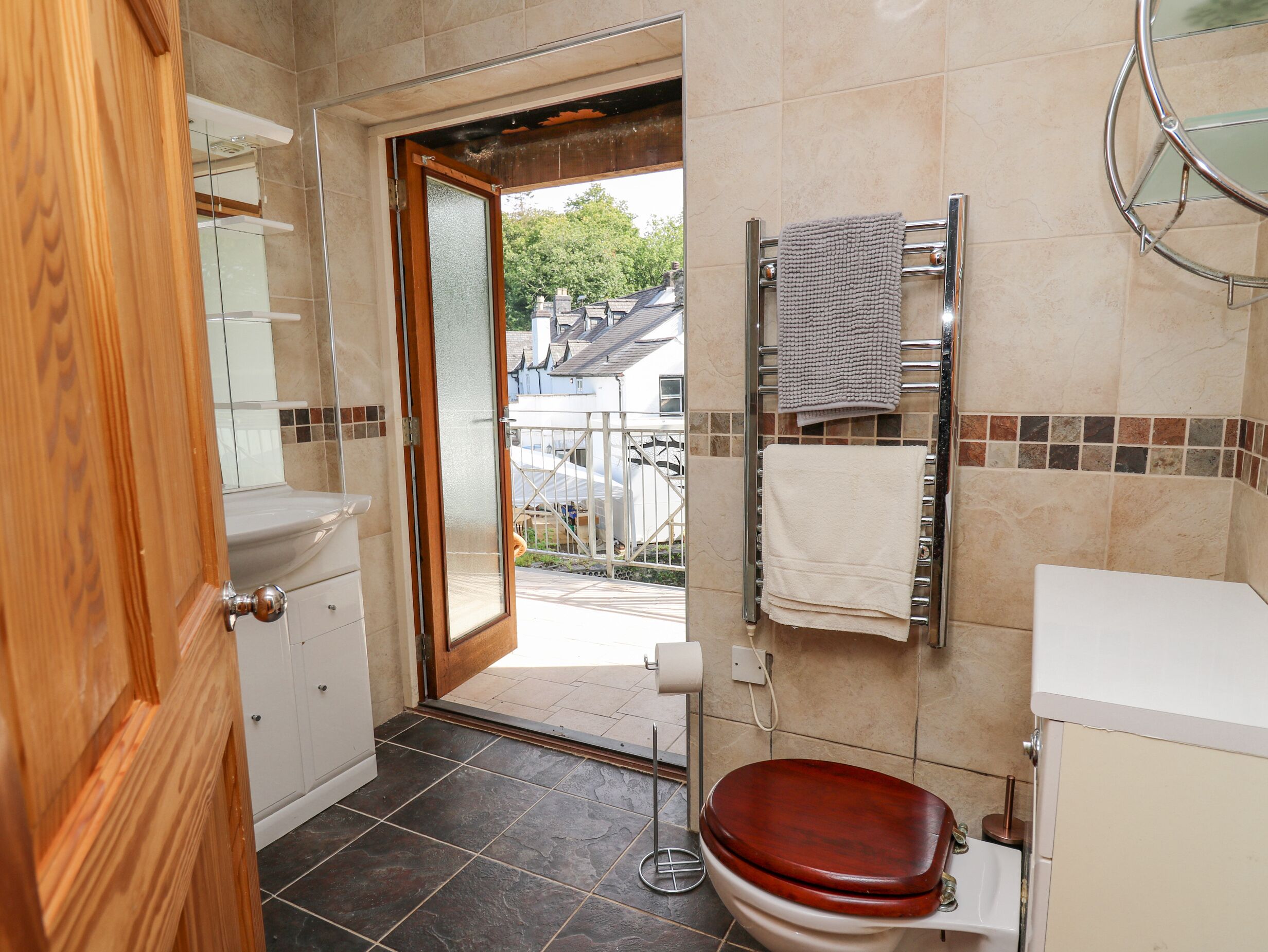 A bathroom with a wooden door, a white sink, a toilet with a wooden seat, a towel rack, and a view of the outside.