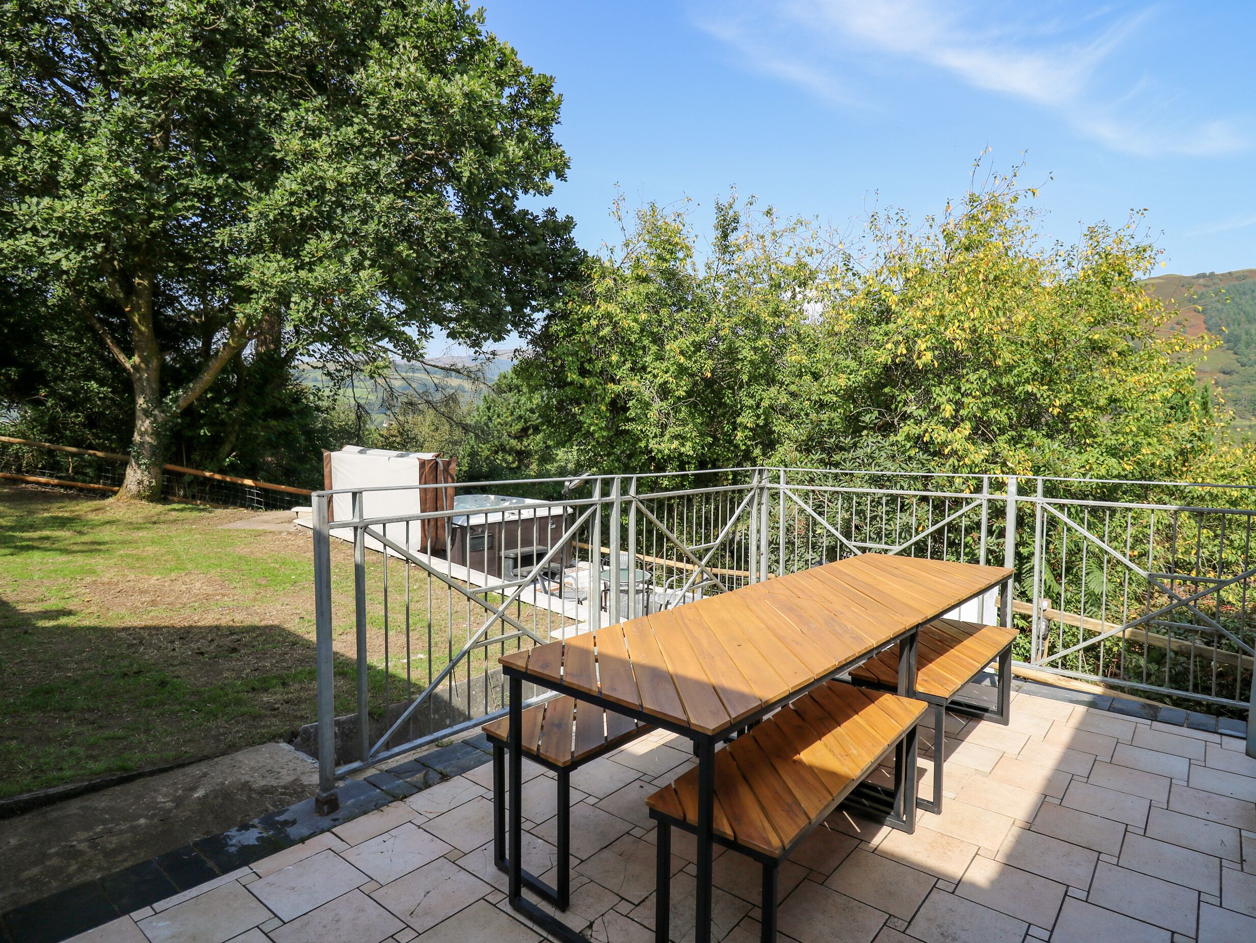An outdoor seating area with a wooden table and benches, surrounded by a metal railing, overlooking a grassy area and trees.