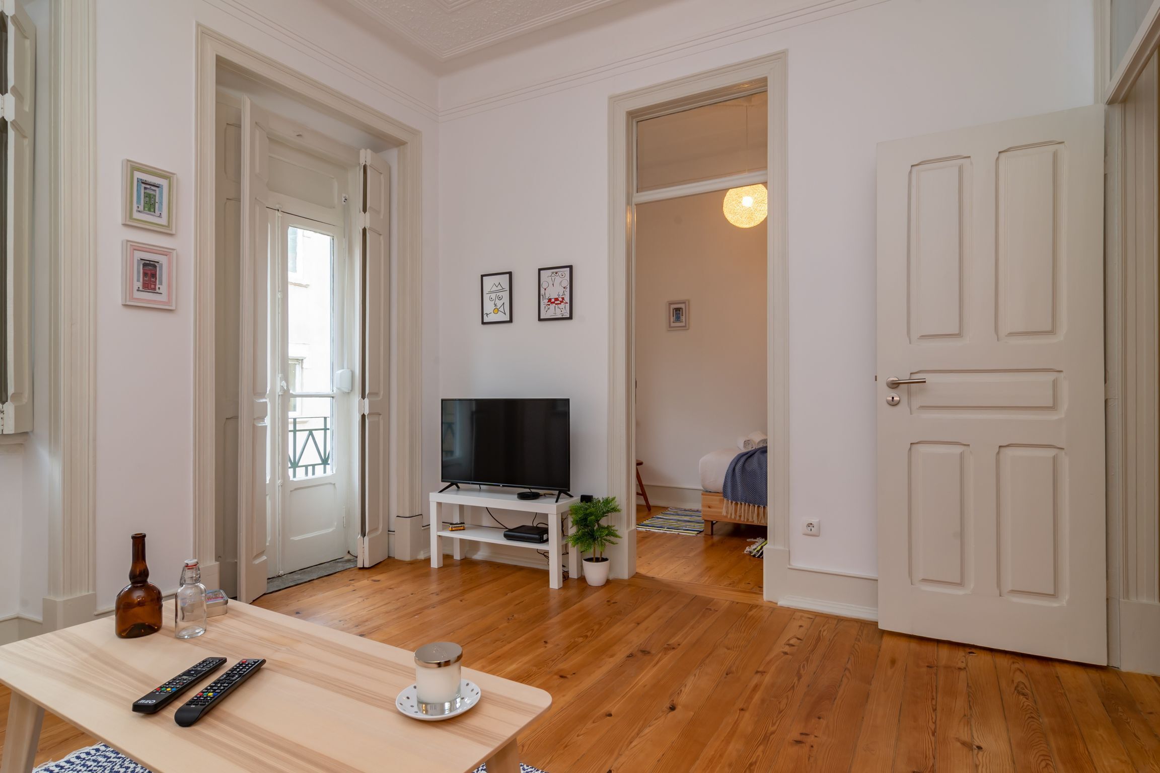 Una sala de estar con piso de madera, un televisor sobre un soporte blanco, un control remoto, una vela y una botella en una mesa.