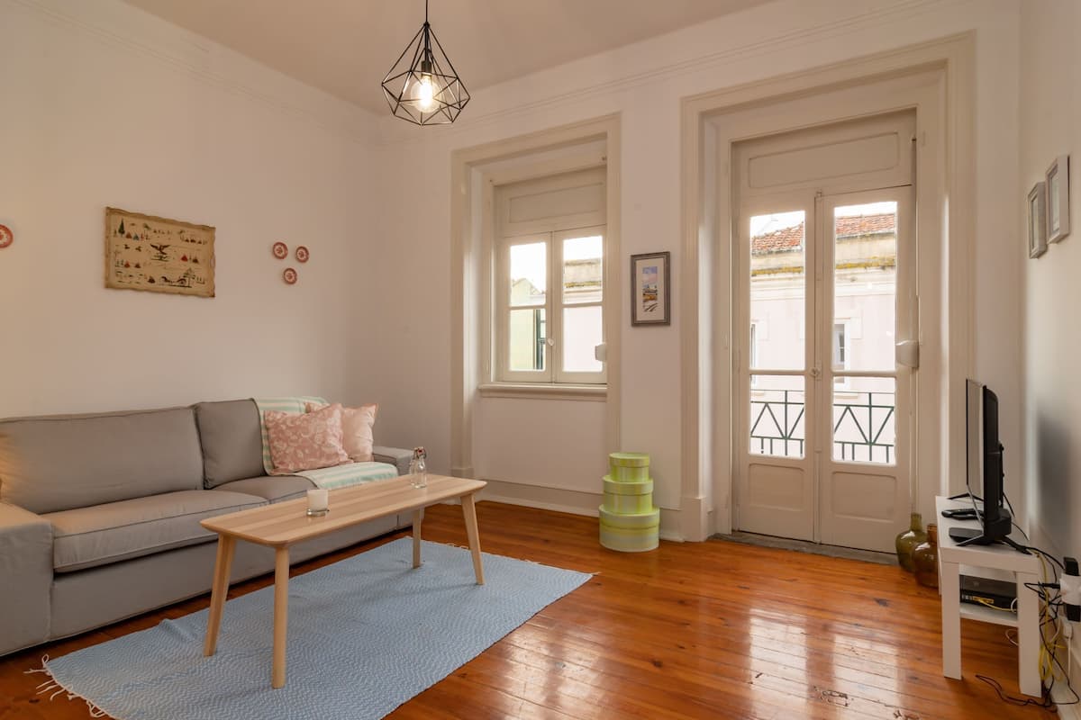 A living room with a sofa, a coffee table, a TV stand, and a window with a view of buildings.