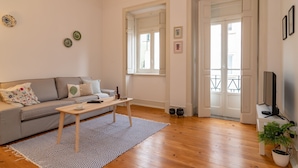 A living room with a sofa, coffee table, and a TV on a stand. There are two windows and a door leading outside.