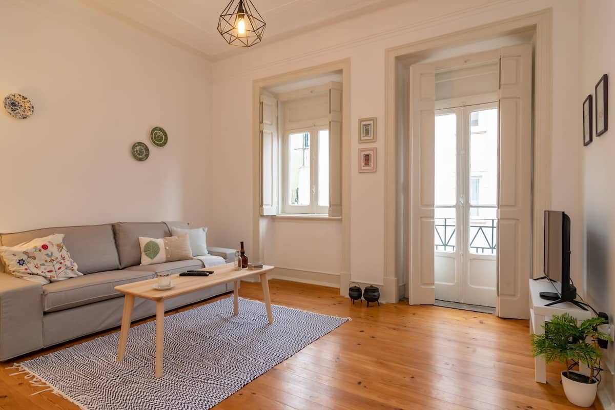 A living room with a sofa, coffee table, and a TV on a stand. There are two windows and a door leading outside.