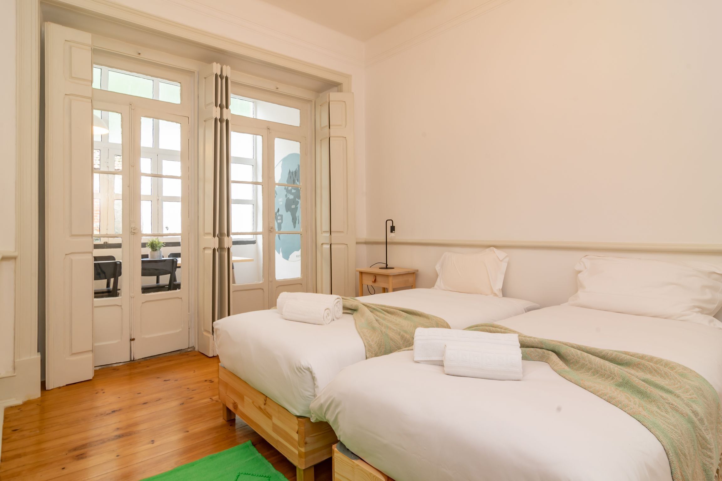 A bedroom with two beds, a wooden floor, a green rug, and a view through a door.