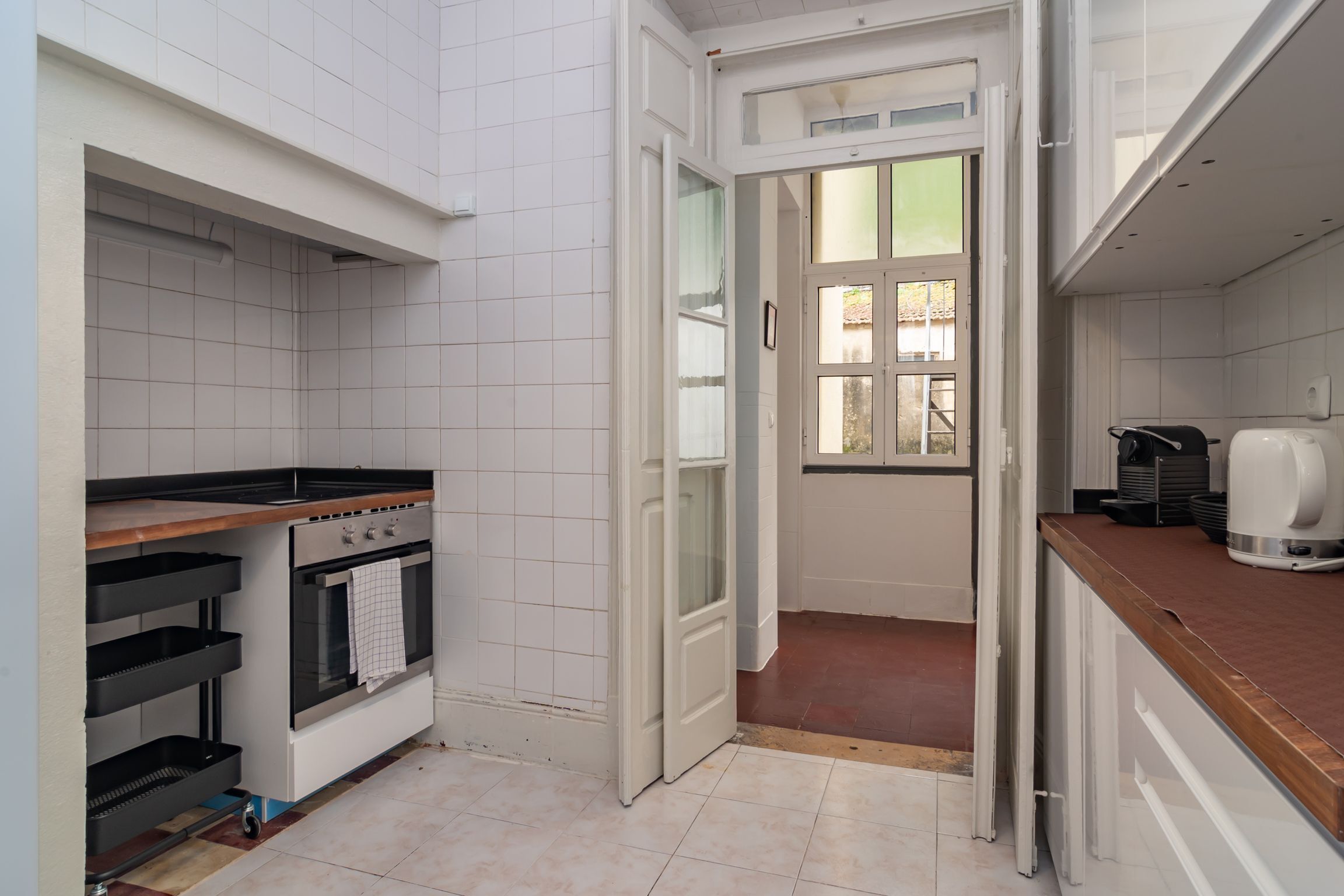 Una cocina compacta con paredes de azulejos blancos, horno empotrado, encimera negra y ventana con vistas.