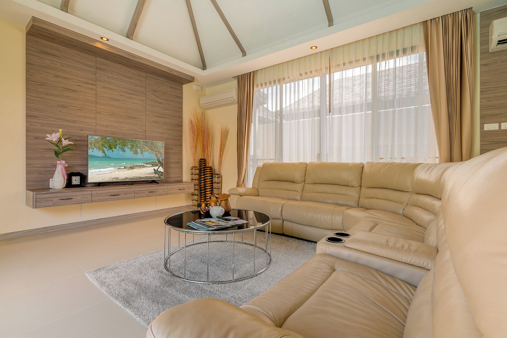 A living room with a beige sofa, a coffee table, a TV on a wooden wall, and a large window with curtains.