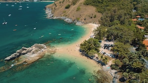 Vista aérea de uma praia com águas cristalinas, afloramentos rochosos e uma extensa linha de árvores.