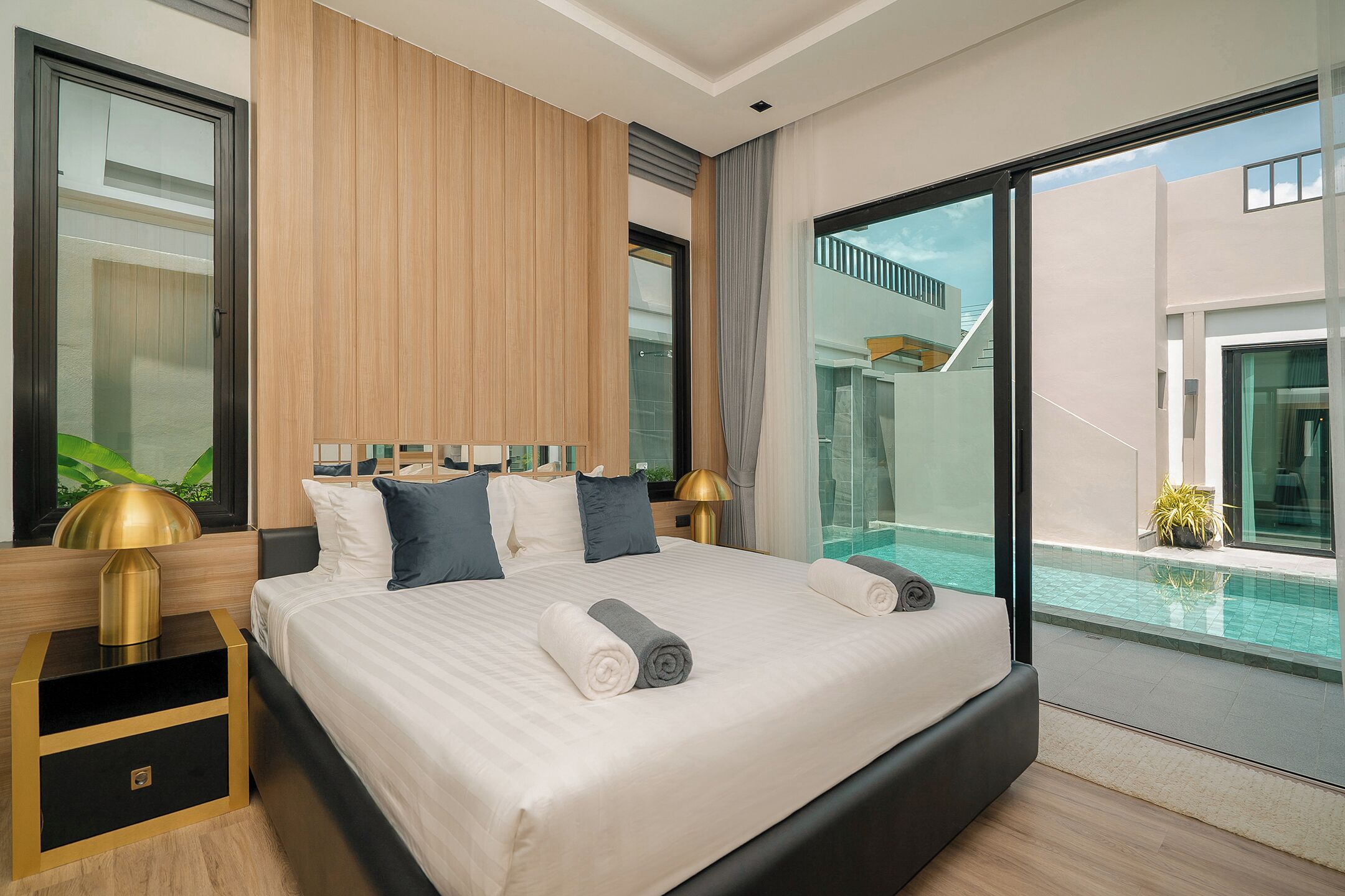 A modern hotel room with a large bed, a nightstand, a lamp, and a view of a swimming pool.