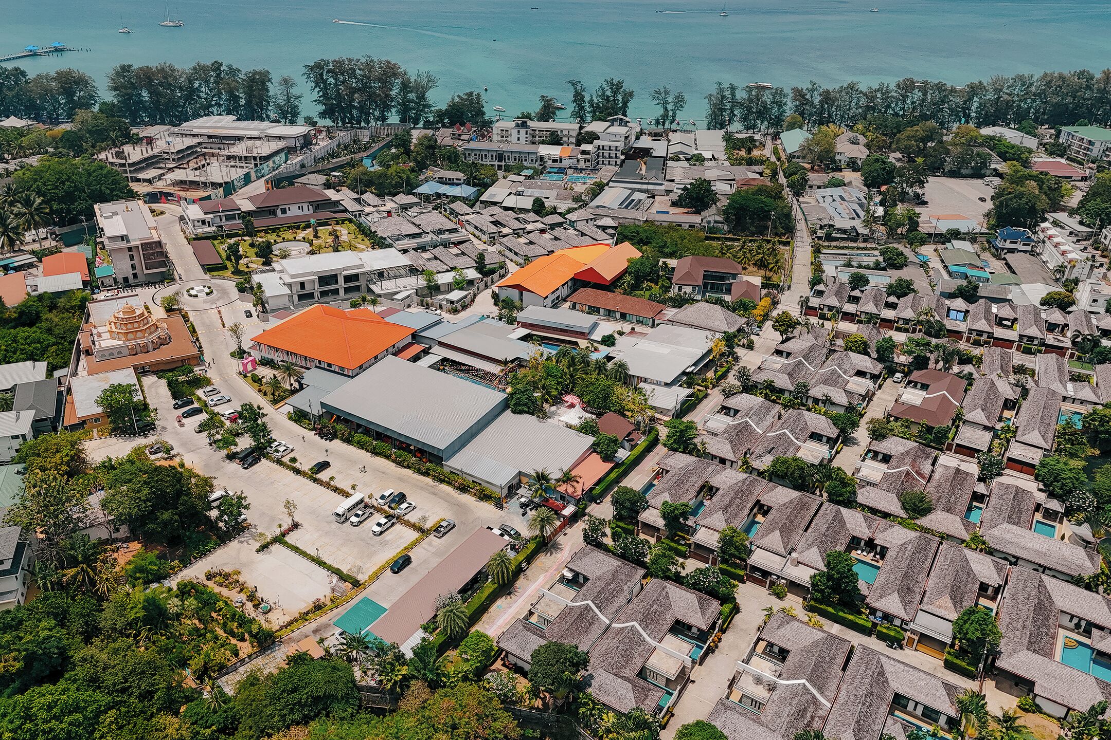 An aerial view of a coastal residential area with a mix of modern and traditional architecture, including a prominent building with a red roof and a large open space with a pool.