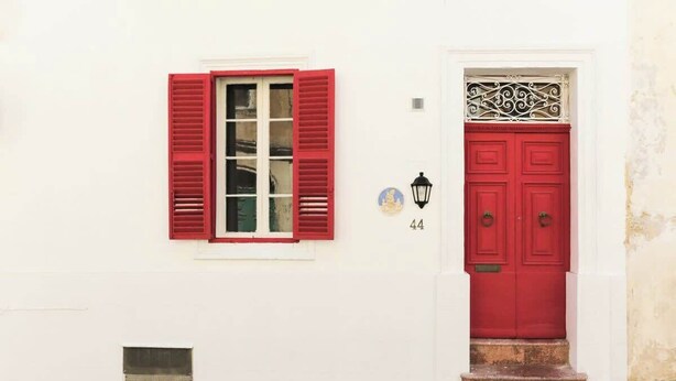 A white building with a red door and window shutters, a small window with a white frame, and a number '44' on the wall.