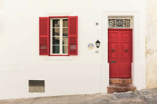 A white building with a red door and window shutters, a small window with a white frame, and a number '44' on the wall.