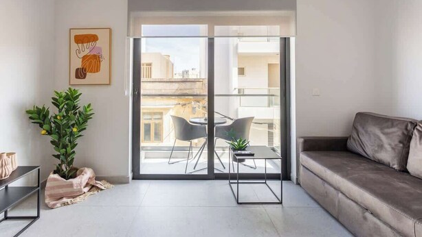 A modern living room with a sofa, a dining table with chairs, a potted plant, and a sliding glass door leading to an outdoor balcony. - FL2 Sliema Cozy 1BR Getaway with Balcony (Sliema)