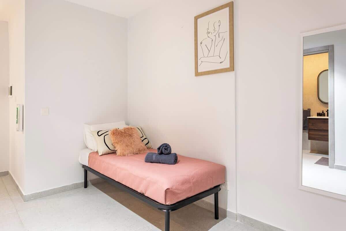 A single bed with a pink bedspread, a fluffy pillow, and a rolled towel. A framed artwork is on the wall above the bed.