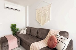 A living room with a brown sofa, a macrame wall hanging, a small plant, and a pink throw blanket.
