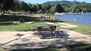 A picnic area with a table and benches, surrounded by a grassy park with a lake and people swimming. - Le Grand Pirou Lumineux Halte Auvergne (Thiers)