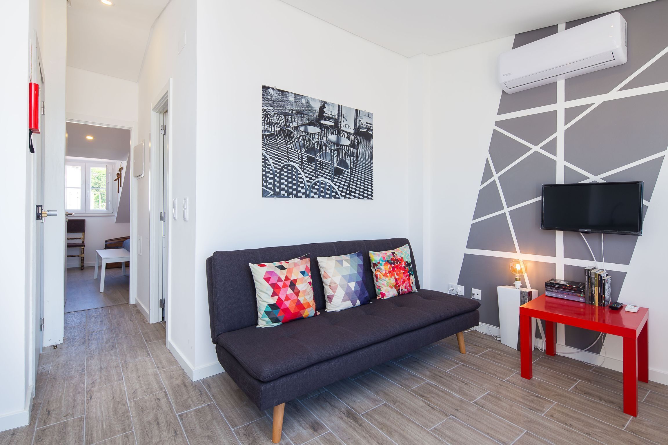 A modern living room with a dark sofa, colorful cushions, a red side table, a mounted TV, and a geometric-patterned wall.