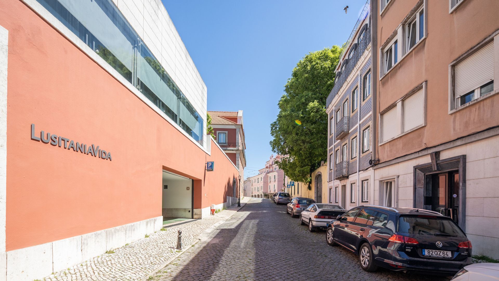 A cobblestone street lined with parked cars and buildings, including a modern building with 'LUSITANIA' signage.