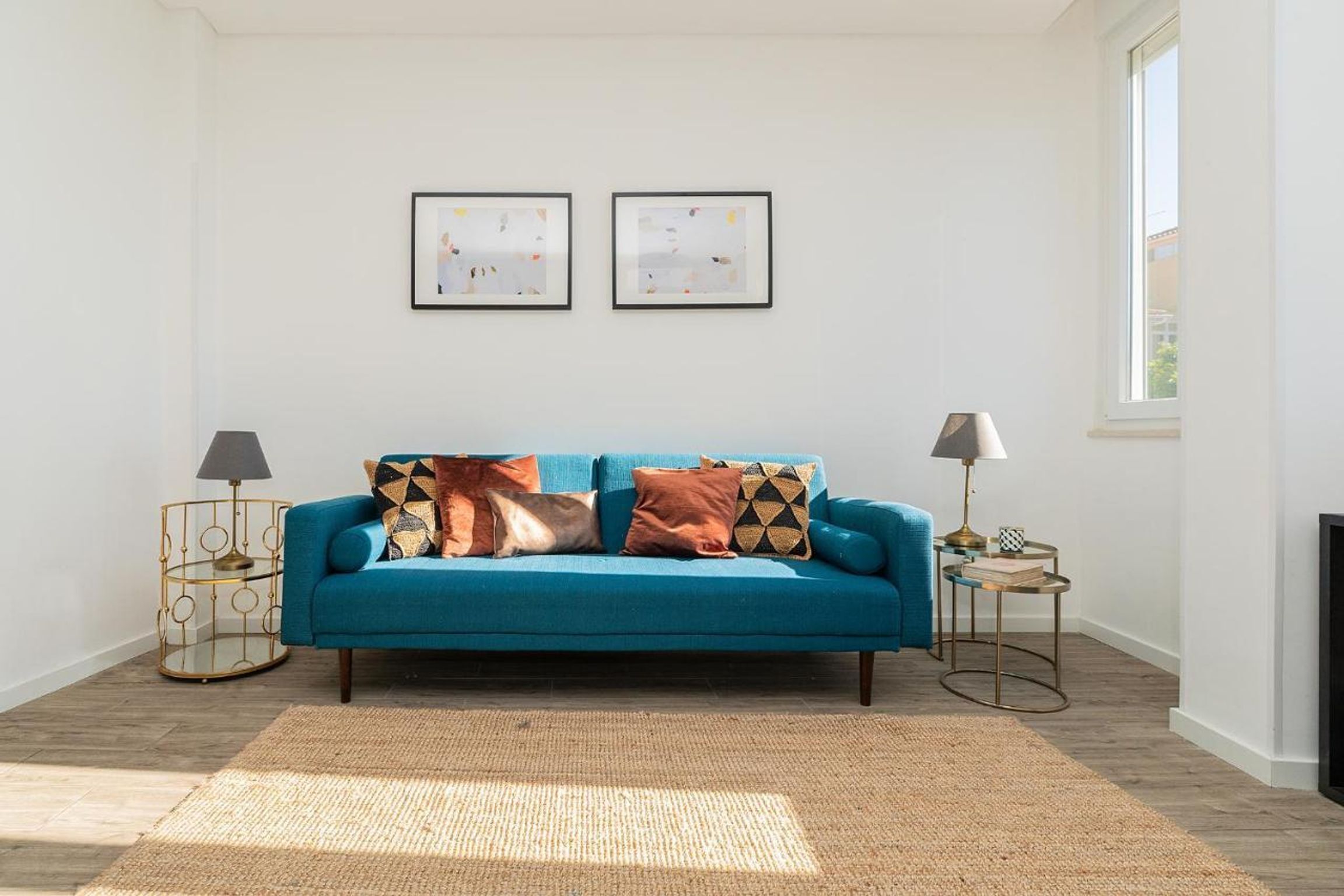 Un salon avec un canapé de couleur turquoise, deux tables d’appoint avec des lampes, un tapis tissé et deux tableaux encadrés accrochés au mur.