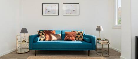 Un salon avec un canapé de couleur turquoise, deux tables d’appoint avec des lampes, un tapis tissé et deux tableaux encadrés accrochés au mur.