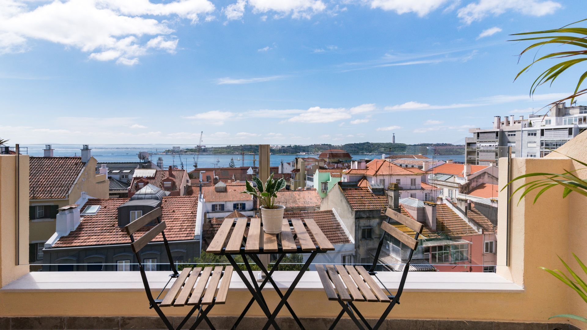 Ein Balkon mit einem Holztisch und Stühlen, der auf eine Küstenstadt mit terrakottafarbenen Dächern und Meerblick blickt.