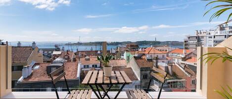 Un balcón con mesa y sillas de madera, con vistas a una ciudad costera de tejados de terracota y al mar.
