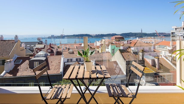 A terrace with a wooden table and chairs, overlooking a coastal cityscape with buildings and a body of water. - Designer Flats in Lapa by LovelyStay (Lisbon)