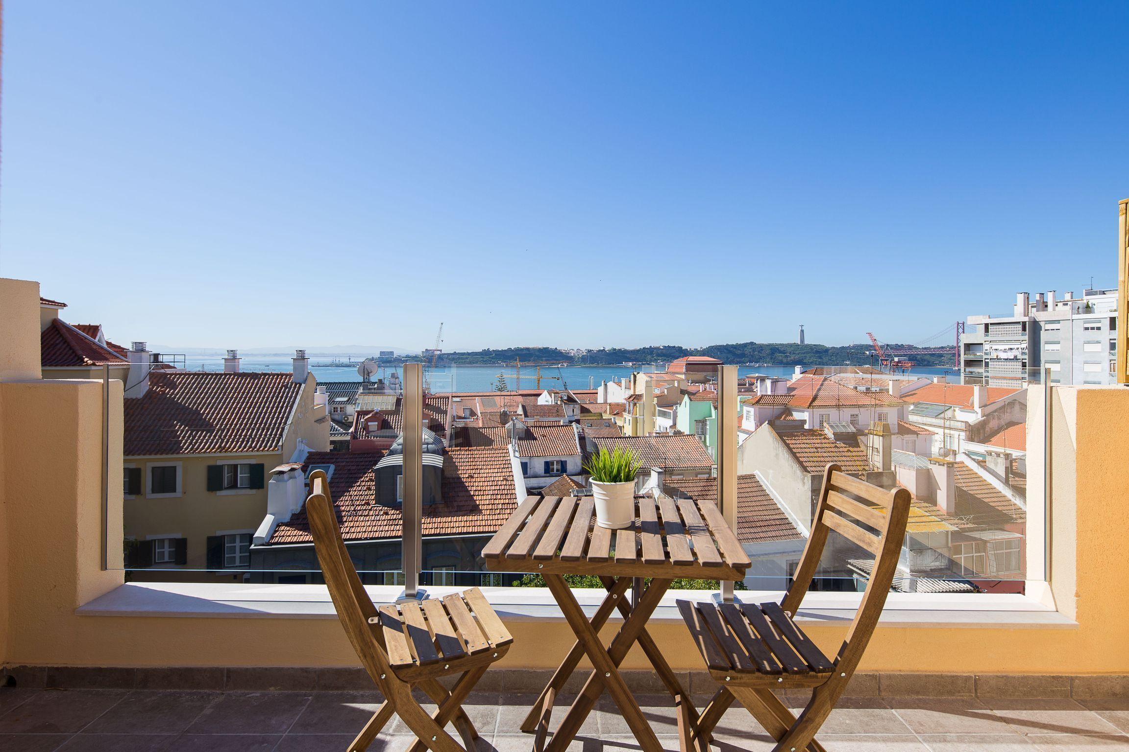A balcony with a wooden table and chairs, overlooking a cityscape with buildings and a body of water.