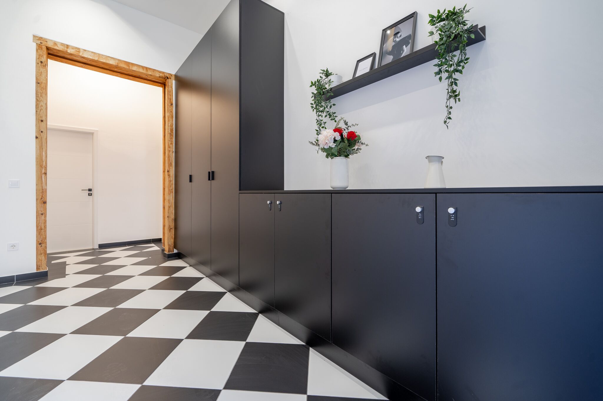 A hallway with a checkered floor, dark cabinets, a wooden door, and a wall-mounted shelf with a plant and a vase.