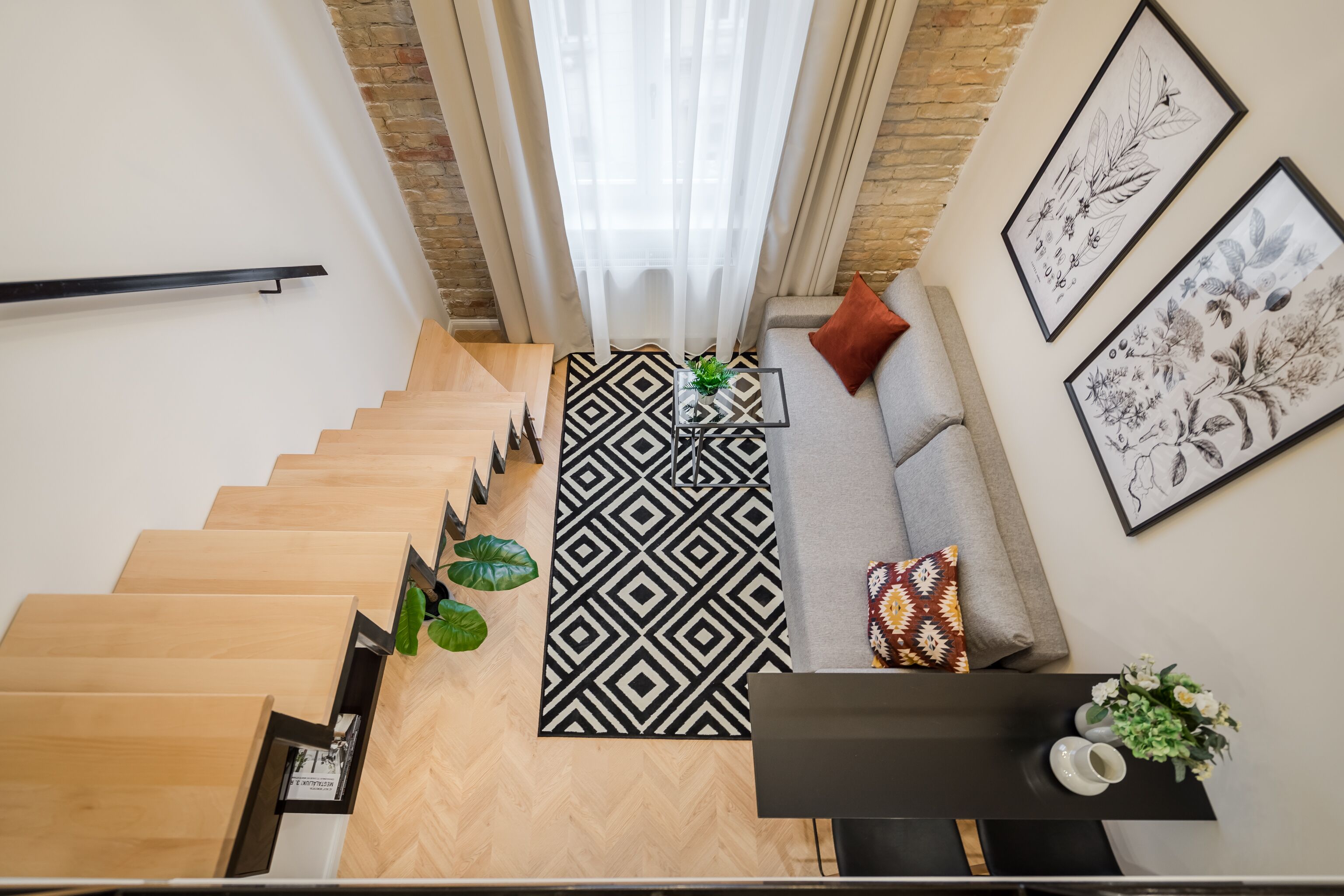 A modern living room with a staircase, a patterned rug, a sofa with cushions, and framed botanical prints on the wall.
