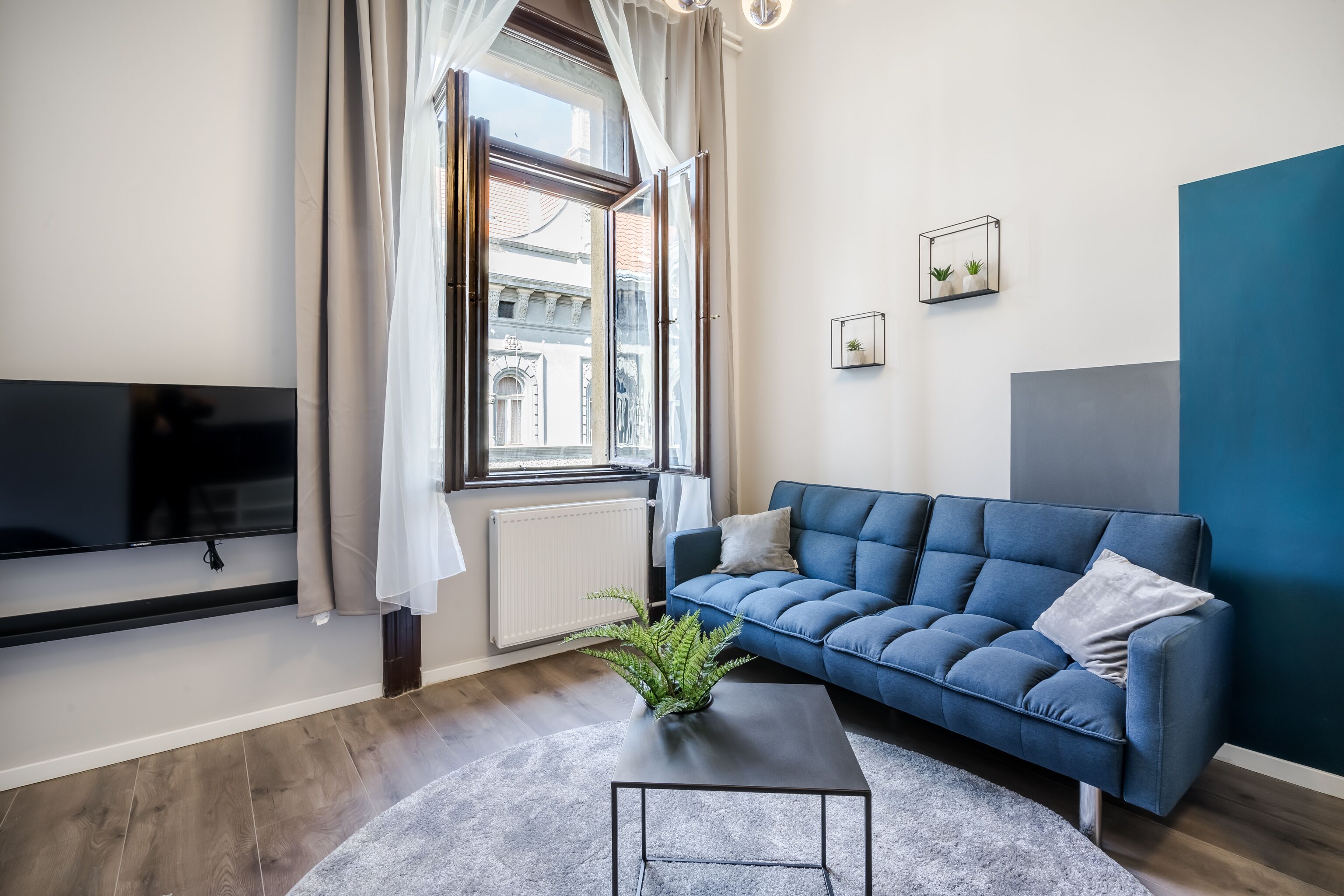 Una sala de estar moderna con un sofá azul, un televisor de pantalla plana, un ventanal con cortinas, un radiador y una pequeña planta sobre una mesa de centro.