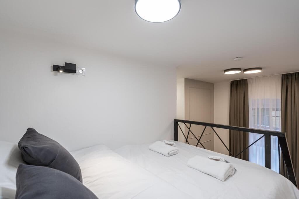 A hotel room with a bed, two pillows, a nightstand, a window with curtains, and a ceiling light.