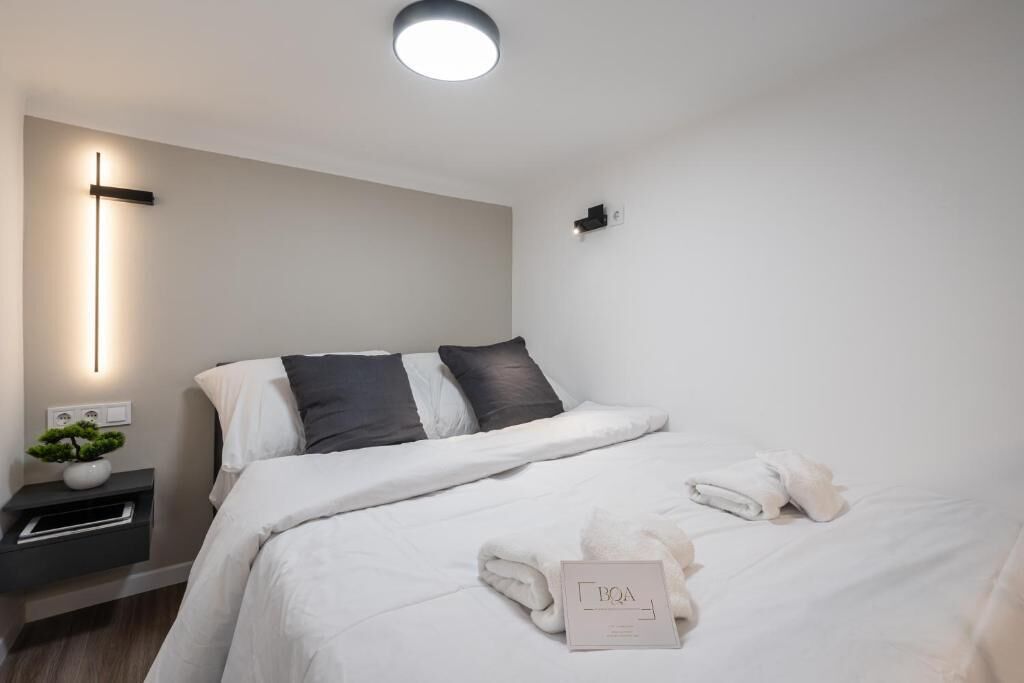 Une chambre d’hôtel moderne avec un lit soigneusement fait, une table de chevet agrémentée d’une plante et un luminaire fixé au mur.