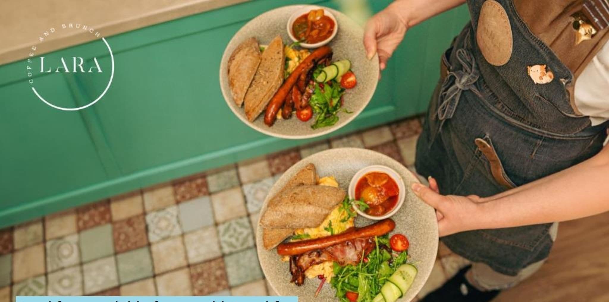 A person holding two plates of food, one with sausages and salad, and the other with eggs and bacon, in a cafe setting.