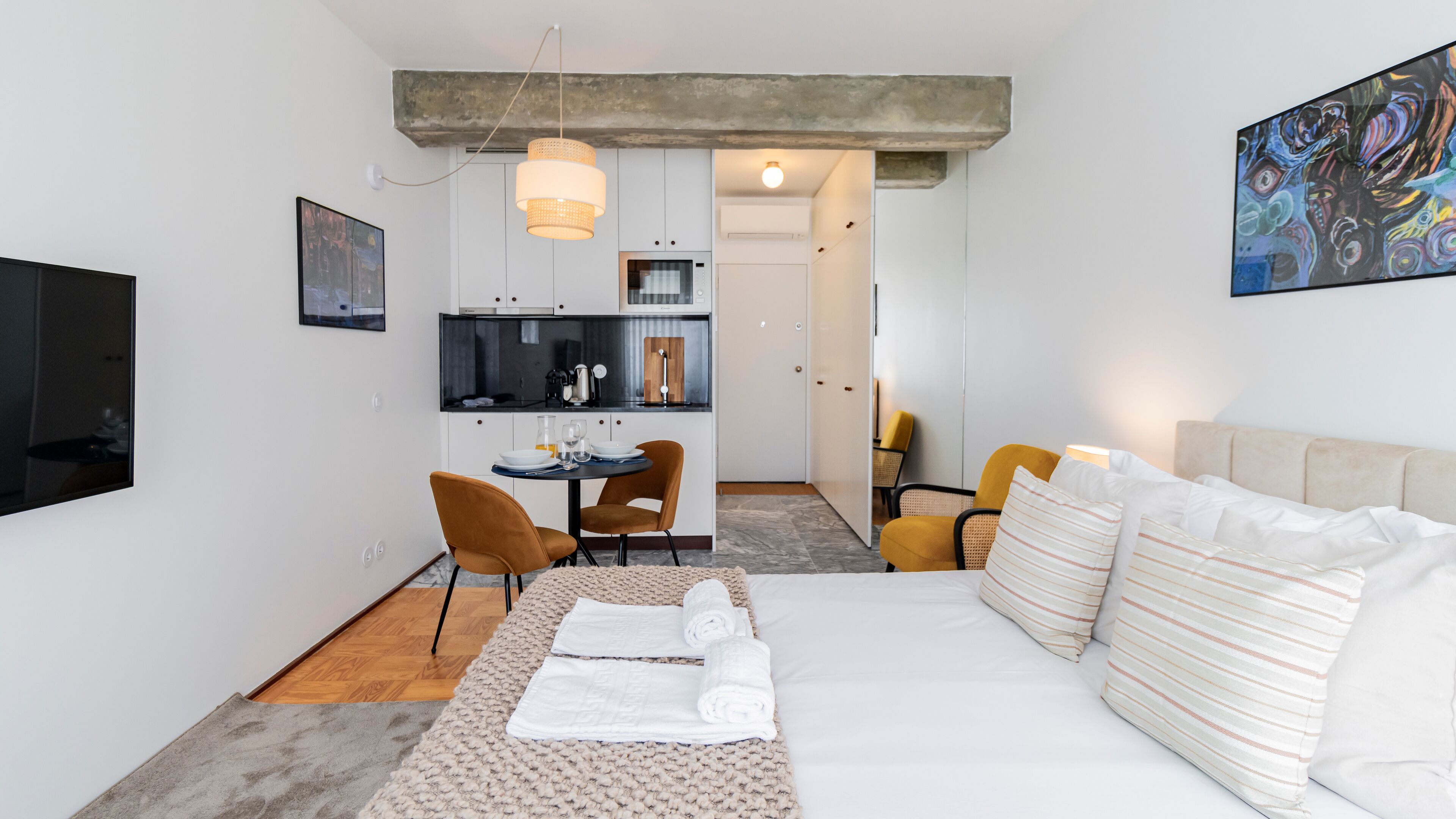 A modern hotel room with a bed, a dining table, chairs, a kitchen area, and a TV.