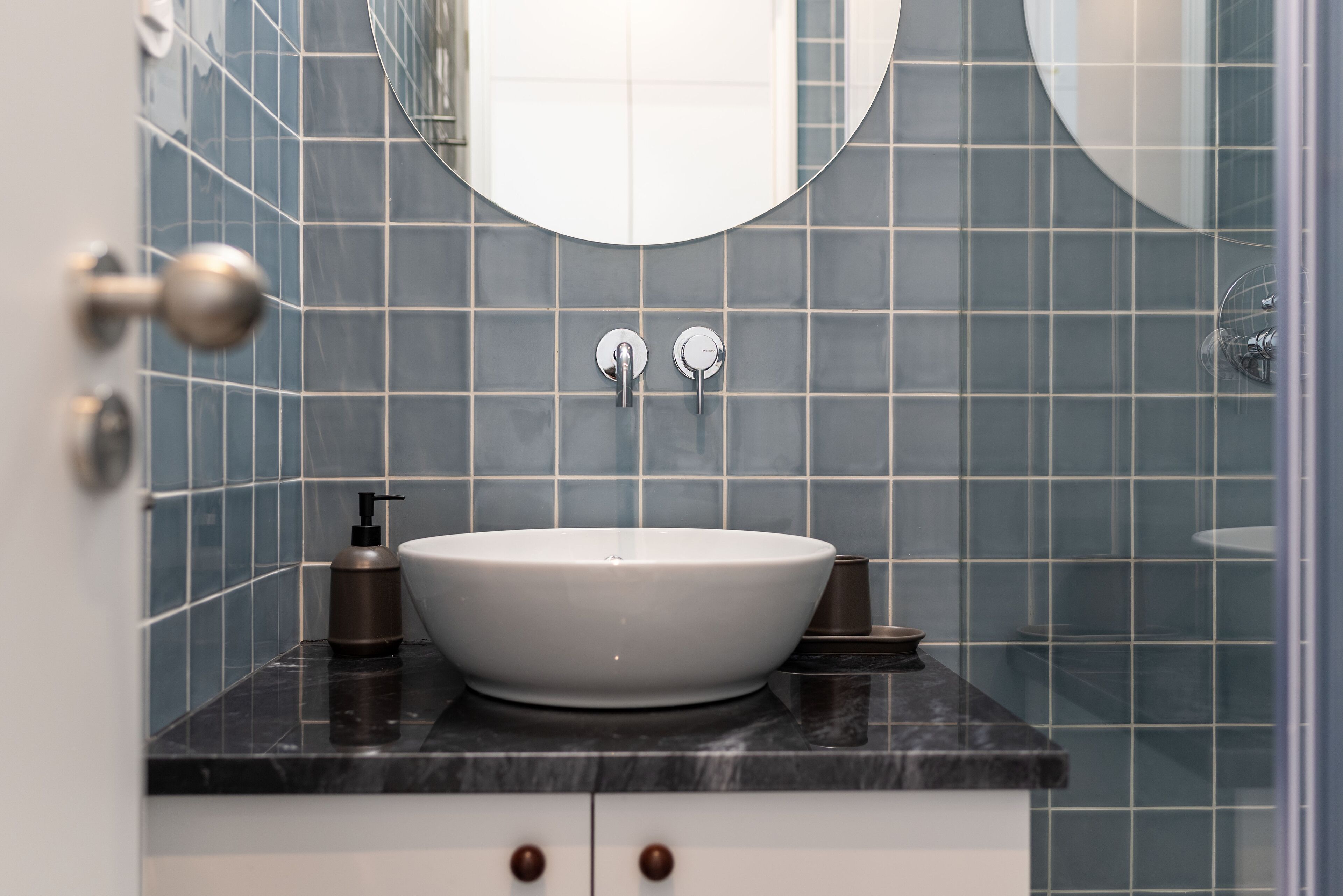 Une salle de bain avec un lavabo blanc, des carreaux foncés et un miroir rond.