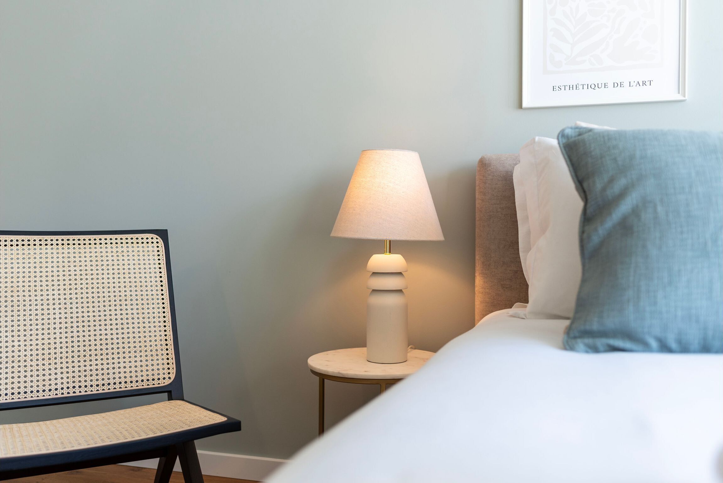 A neatly arranged hotel room with a bed, a small round table with a lamp, a wicker chair, and a framed artwork on the wall.