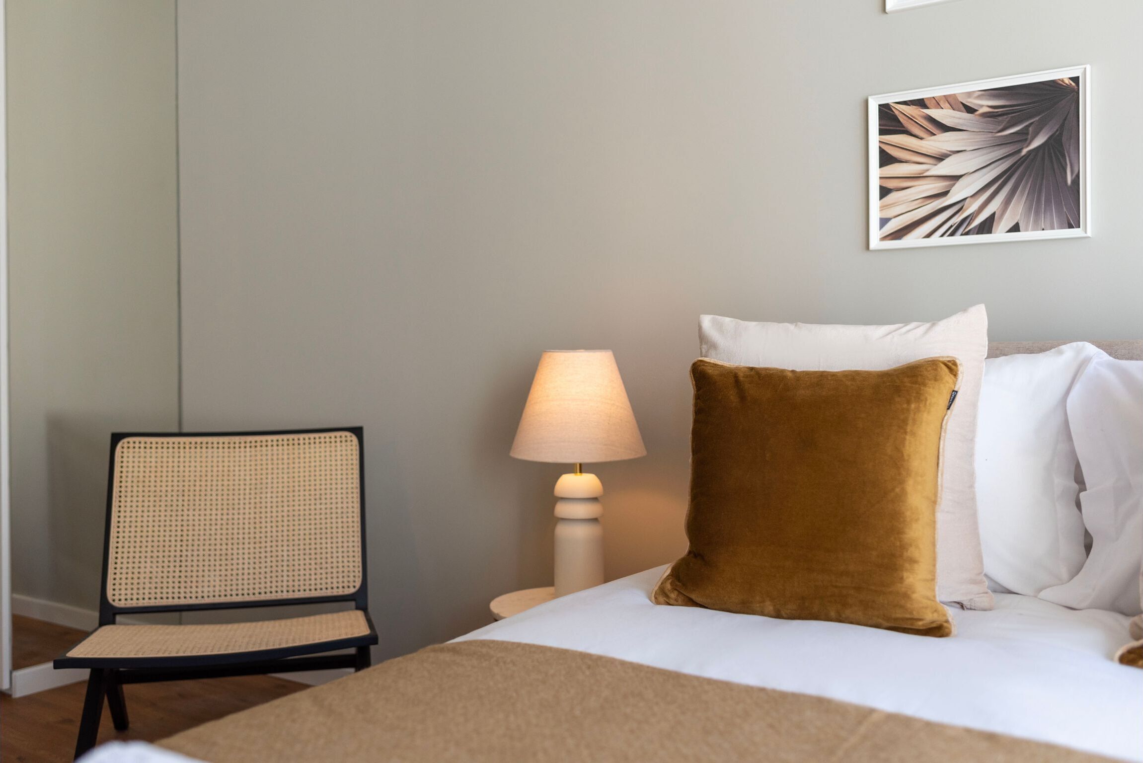 A neatly made bed with a brown pillow, a white lamp, and a framed leaf artwork on the wall.